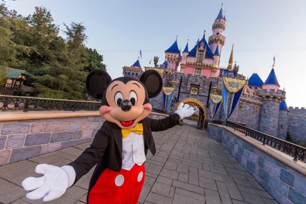 Image of Mickey Mouse with hands open, in a welcoming gesture, in front of Sleeping Beauty Castle at Disneyland Resort.