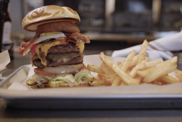 Image of a three patty burger topped with bacon, onion rings, tomatoes, lettuce, and pickles. Burger is served with a side of french fries on top of a metal cookie sheet.