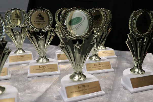 Image of a steel-looking table with nine gold and white trophies placed on top. The trophies are displayed in no particular order on the top and feature the logo of the Anaheim Convention Center and a frisbee/disc icon.