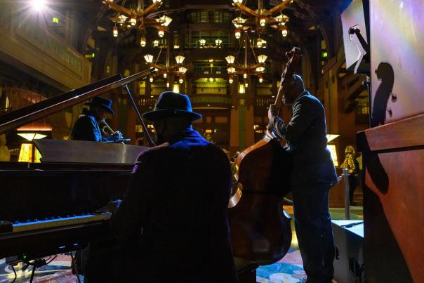 Image of three musicians in a dim-light room. One gentleman is setting, playing a piano. Another gentleman is standing up playing a stand-up cello or bass. The last gentleman in the photo is standing playing a horn or saxophone.