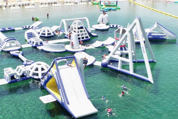 Image of water park located in Anaheim, California. Image shows inflatable obstacles placed inside a large pool of water with people swimming and climbing on each obstacle.