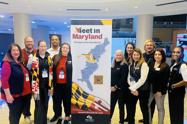 Group photo of Maryland Representatives surrounding the Meet in Maryland Banner (which is a Maryland Flag at bottom, the wordwmark, and east coast US map with a QR Code).