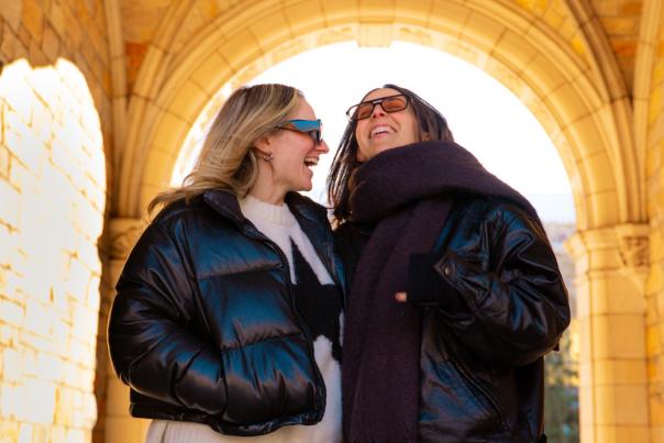 Two women wearing jackets and sunglasses are laughing and smiling as they walk underneath a stone archway.