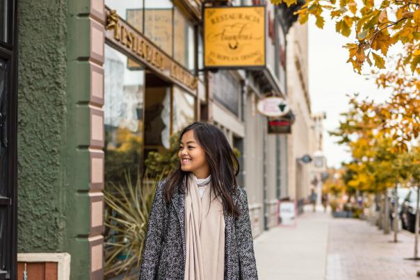 Woman walks down sidewalk and smiles as she looks into storefronts along the way. Trees line the sidewalk with yellow and orange leaves.
