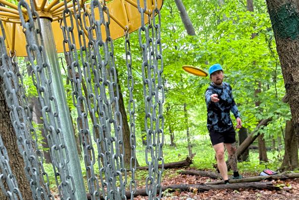 Man throws disc golf frisbee towards camea, which sits behind the hole in heavily wooded and shaded area