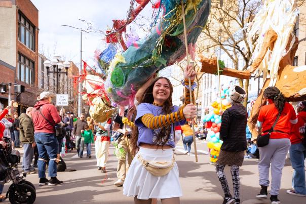 Young woman smiles as she helps support a large puppet with a wooden post during a street festival parade.