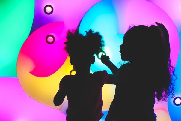 The sillouette of two young girls playing in front of a colorful light wall at the Thinkery.