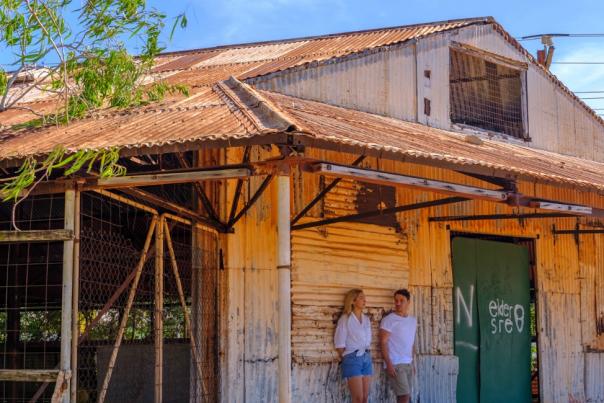 A couple stands against the wall of an old corrugated iron building overlooking Roebuck Bay