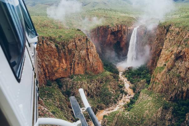 The foreground shows the step rail of the helicopter where this image is taken and one side of the helicopter windows. In the far distance Revolver Falls waterfall spills water over a rock face into a muddy rushing creek below. There is mist coming off the waterfall and lush vegetation all around