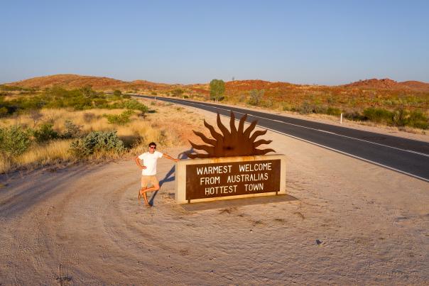 Man standing at the Marble Bar welcome sign which reads "Warmest welcome from Australia's hottest town."