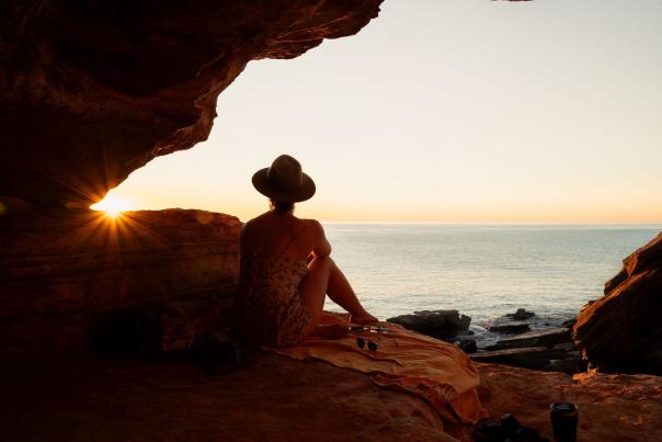 A woman wearing a hat and sitting on rocks at Gantheaume Point, silhouetted by the setting sun.