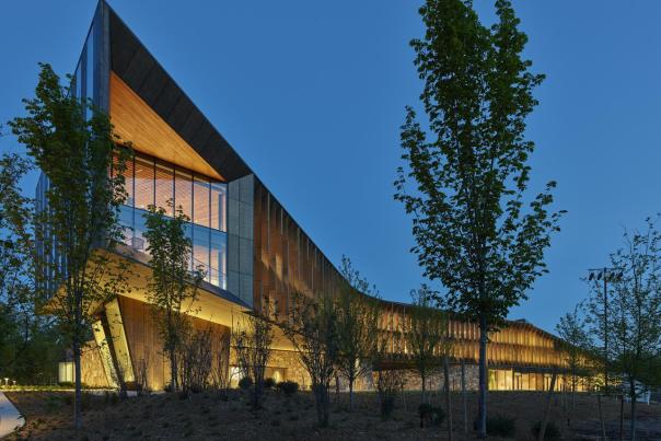 Exterior view of the Heartland Whole Health Institute at dusk, featuring angular modern architecture, warm interior lighting, and surrounding trees.