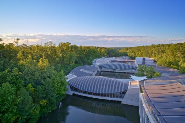 Aerial view of modern architectural structures nestled among lush greenery, reflecting on a serene waterway under a clear blue sky.