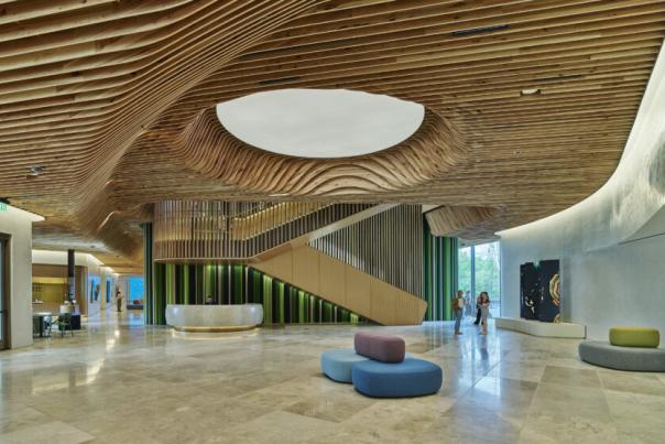 A grand, modern lobby featuring a wavy slatted wood ceiling with a circular cutout, a large wooden staircase, and a curved reception desk with a green textured front. Light-colored stone floors, colorful ottomans, and large windows with people and trees complete the scene.