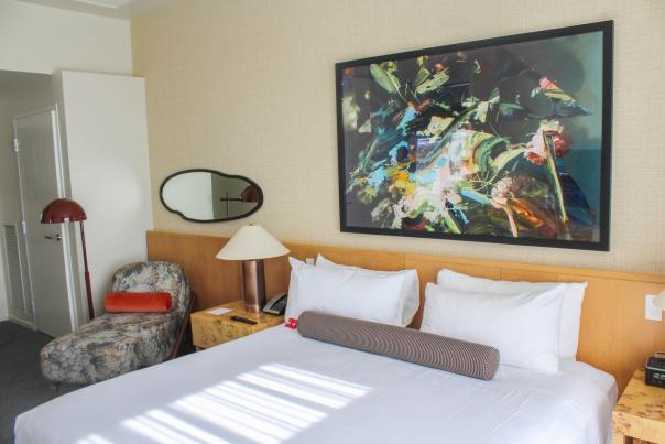 A cozy hotel room featuring a queen bed with white linens, a colorful abstract painting, a lamp, and a patterned armchair.