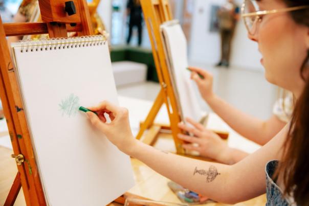A woman draws on a sketchpad with a green crayon at an art workshop. Easels and a bright, modern studio setting are visible.