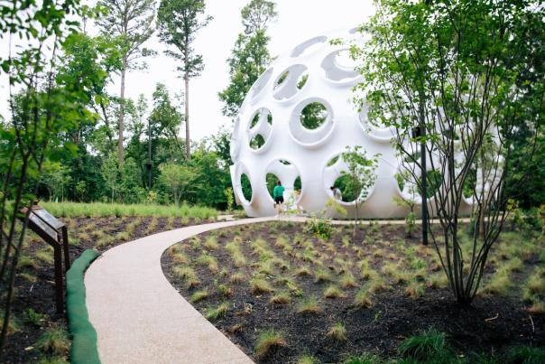 A winding path leads through lush greenery to a modern, spherical structure with circular openings, surrounded by trees and plants.
