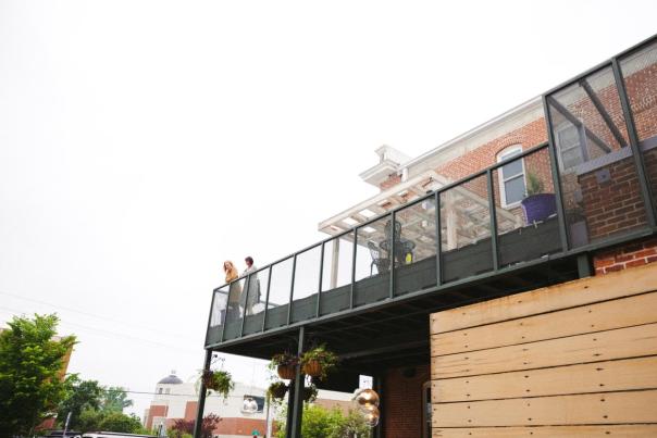 A modern balcony with two people standing, overlooking a vibrant street scene. Lush greenery and decorative elements enhance the inviting atmosphere.