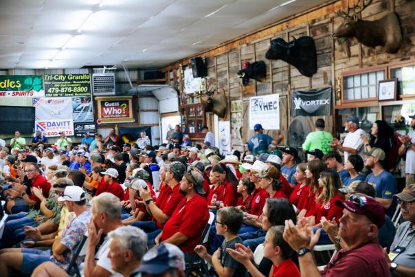 A crowded indoor venue with attendees in red shirts, clapping and engaging. The rustic walls feature mounted animal heads and various banners.