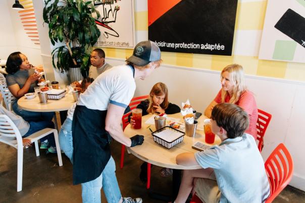 A casual dining scene featuring a waiter serving a family at a round table, surrounded by vibrant wall art and greenery.