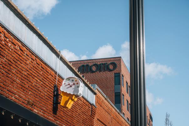 Brick building with a large yellow ice cream cone sign featuring white ice cream and colorful sprinkles; the Motto hotel is in the background, with a tall black pole and blue sky overhead.