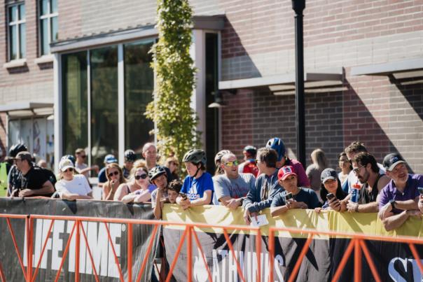 Spectators of various ages stand behind a barricade at Big Sugar Bicycle Classic, in front of a brick building with large windows. Some wear helmets and sports attire, while others are casually dressed. Banners on the barricade display the word 'LIFETIME' as the crowd watches, takes photos, and chats.