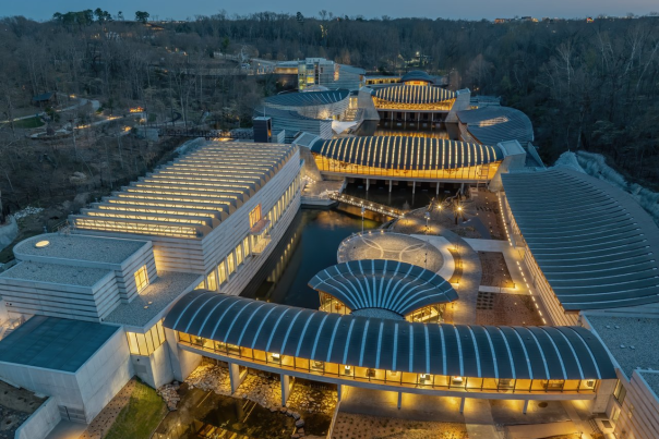 Aerial view of a modern architectural complex with flowing lines, illuminated pathways, and surrounding greenery at dusk. The design features multiple interconnected buildings and water elements.