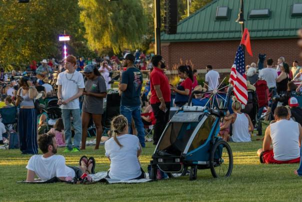A lively outdoor gathering with diverse groups of people enjoying a sunny day on a grassy field, some seated and others standing. A stroller with an American flag is visible, adding to the festive atmosphere.