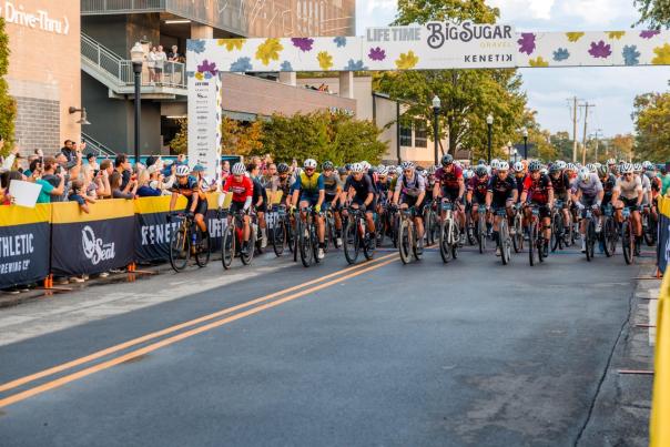 A group of cyclists races under a colorful archway at the Big Sugar Gravel event, with spectators cheering along the roadside.