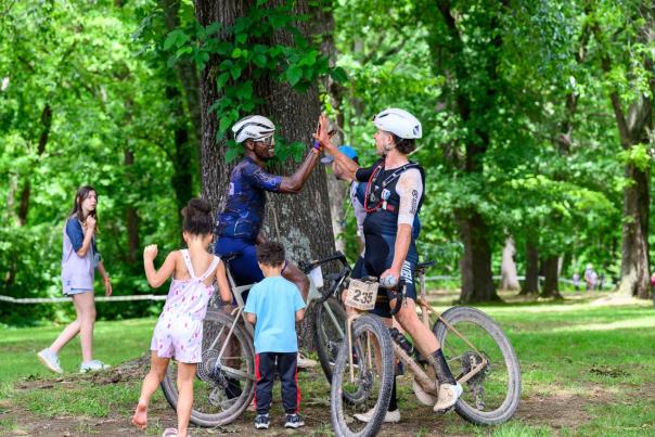 Two mountain bikers in helmets share a high-five beside their bikes, surrounded by children playing in a lush green park.