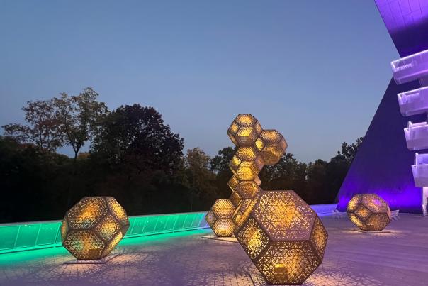 Illuminated geometric sculptures in a modern outdoor space at dusk, surrounded by trees and vibrant lighting, creating a captivating atmosphere.