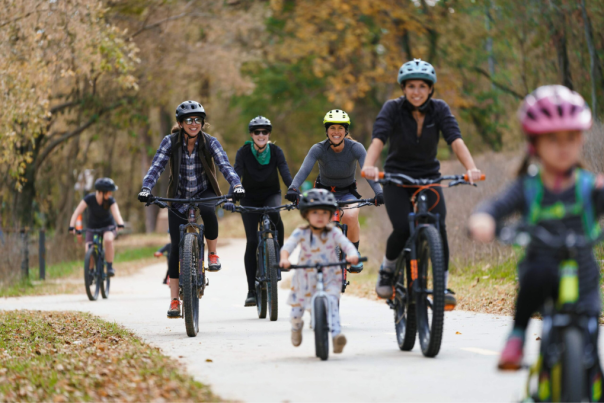 A mix of women and children riding mountain bikes cruise on a paved trail in colorful bike helmets.