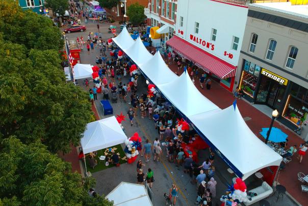 An aerial view of the Bentonville Square with green trees in the foreground and pedestrians strolling among vendor tents.