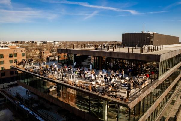 A rooftop bustling with people enjoying an outdoor space, surrounded by modern buildings and a clear blue sky. Ledger, Bentonville.