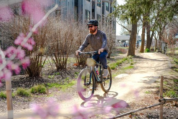 A man rides a mountain bike along a dirt path surrounded by budding trees and blooming flowers, with modern buildings in the background.