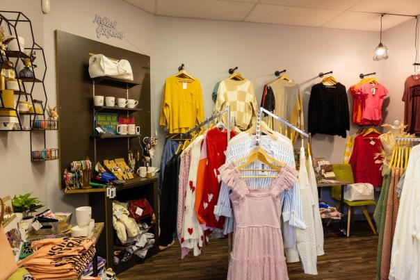 A cozy boutique interior featuring colorful clothing racks, accessories, and decorative items. Shelves display mugs and gifts, creating a vibrant shopping atmosphere.