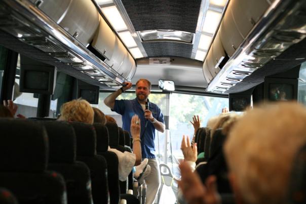 A tour guide speaks to a group on a bus, with passengers raising their hands. The interior features overhead storage and screens.