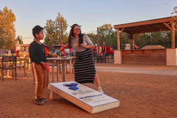 A woman in sunglasses plays cornhole with a boy in a casual outdoor setting, surrounded by tables and trees at sunset.