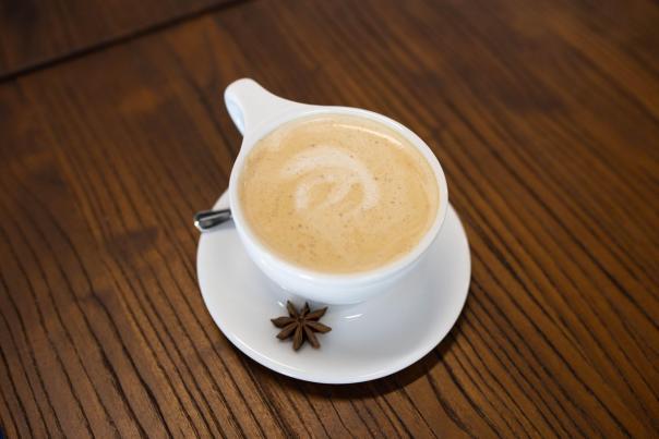 A creamy latte in a white cup sits on a round saucer, accompanied by a star anise, on a wooden table. The latte features a delicate foam design.