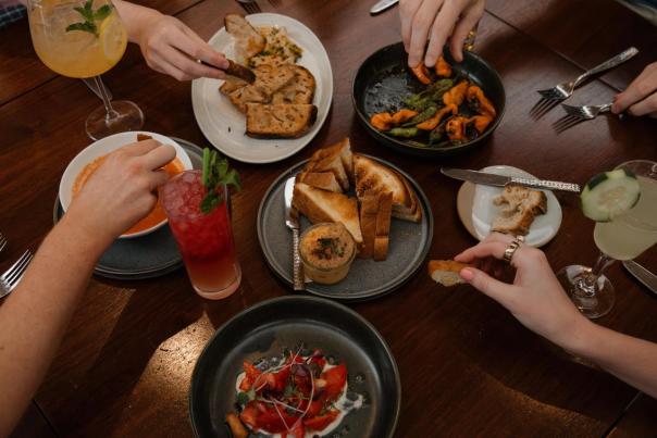 A table set with various dishes, including toast, salads, and drinks. Hands reach for food, creating a lively dining atmosphere.