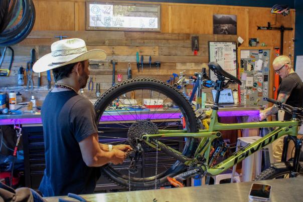 A bike mechanic adjusts a green mountain bike in a workshop filled with tools. A second worker is seen in the background, focused on repairs.