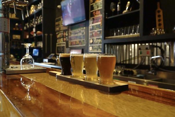 A selection of craft beers in varying shades on a flight board at The Bentonville Taproom, with a view of the bar and brewing equipment in the background.