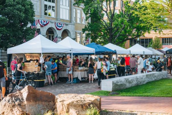 A bustling outdoor market with white and blue tents, vendors selling goods, and a crowd of people browsing. Green trees and historic downtown Bentonville buildings in the background.