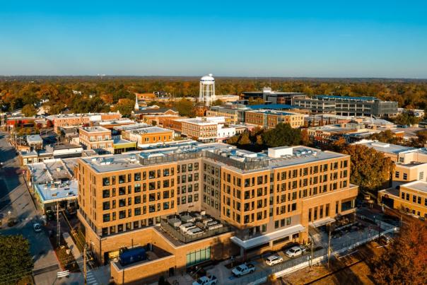 Aerial view of a modern hotel in a vibrant downtown area, surrounded by historic buildings and lush trees under a clear blue sky.