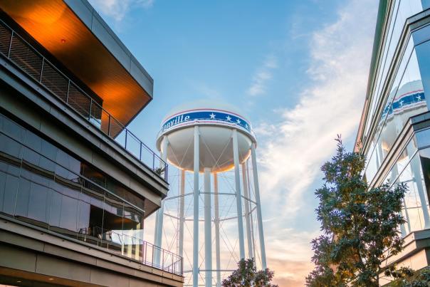A water tower with "Bentonville" and stars stands between modern buildings under a blue sky with wispy clouds, creating an urban scene.