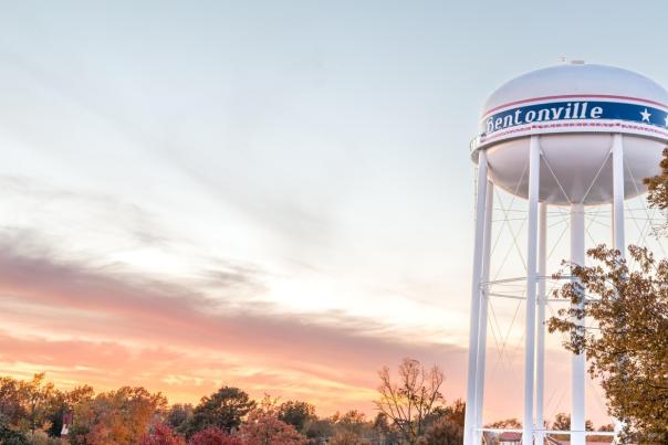 Water tower with "Bentonville" signage against a  sunset sky, surrounded by trees in autumn colors. A serene local scene.
