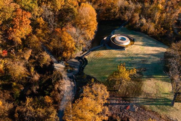 Aerial view of James Turell SkySpace: The Way of Color surrounded by autumn foliage, with winding paths and grassy areas nearby.