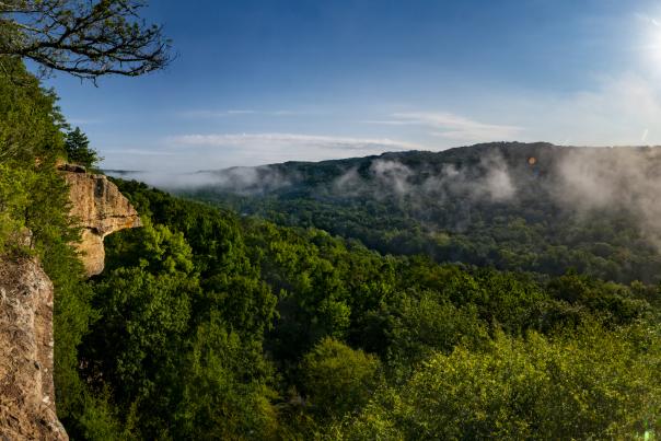 Panoramic view from Yellow Rock Trail at Devil’s Den State Park in Arkansas, featuring a rocky cliff overlook above a forested valley with morning fog drifting through the hills beneath a bright blue sky.