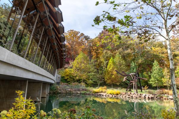 An image of one exterior wall at Crystal Bridges Museum of American Art next to a body of water and in front of fall leaves