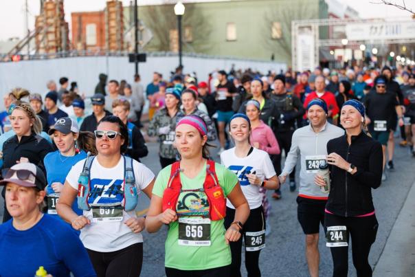 A large group of runners in athletic gear participate in a race, with a mix of men and women, on a city street. The atmosphere is energetic and vibrant.
